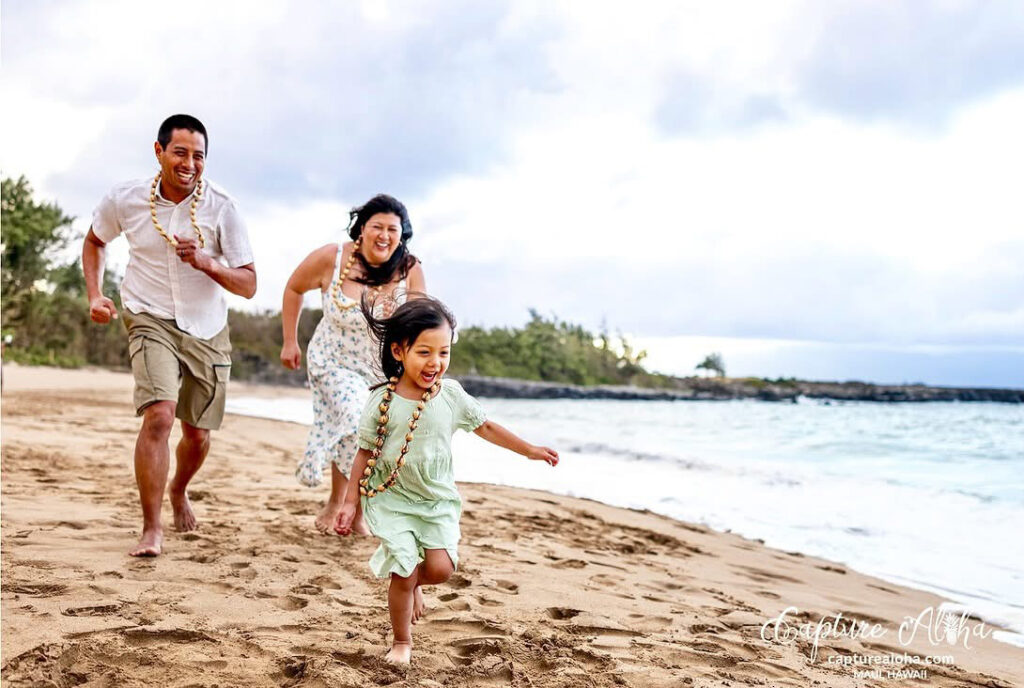 Maui family photography – parents chase their laughing daughter along the shoreline, leis swinging, candid joy on a sandy Maui beach at golden hour.