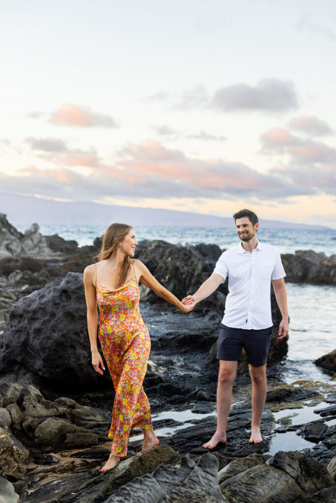 Maui engagement photos at Ironwoods (Oneloa) Beach – couple holding hands along the shoreline, rugged lava rocks and pastel sky in West Maui.