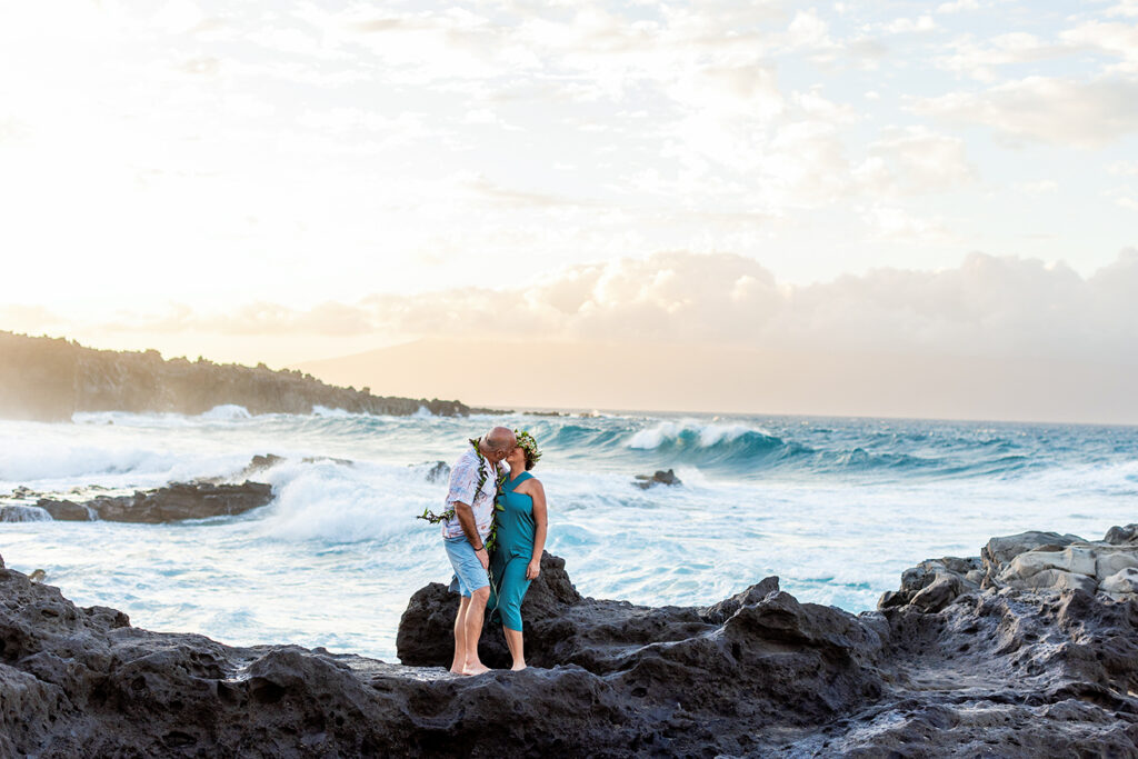 Couple sharing a kiss on the black lava rocks during a Maui couple photo session at Ironwoods Beach with waves behind them.