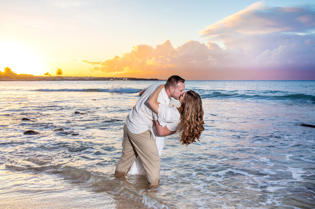 Playful Maui couple photo session, man dipping his partner in the ocean during sunset with colorful skies.