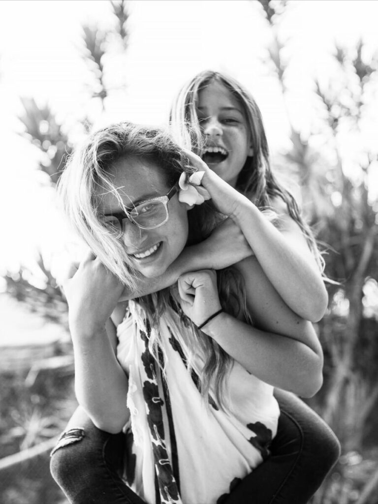 Maui lifestyle photography – mother and daughter laugh in a playful piggyback, tropical foliage bokeh and bright light for a natural, candid portrait in Maui.