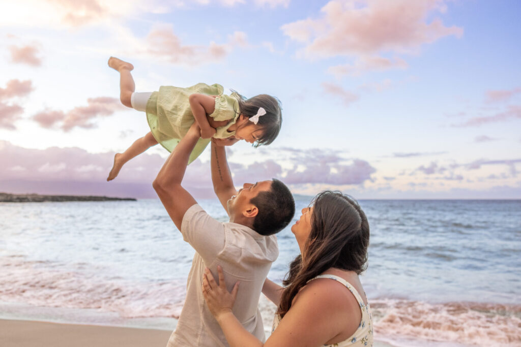 Maui beach portraits – dad lifts daughter into the pastel sky while mom looks on, gentle waves and pink clouds at sunset creating a dreamy Maui moment.