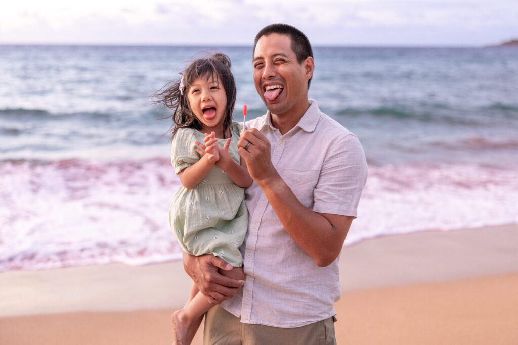 Father and daughter laughing with lollipops by the ocean during Maui family photography at DT Fleming Beach.