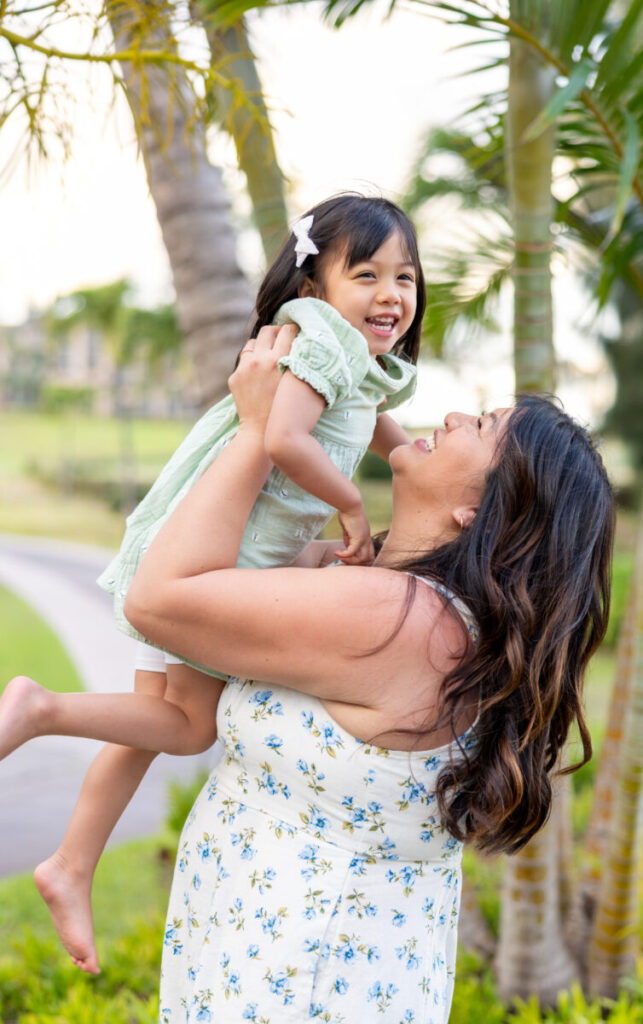 Mother lifting daughter in the air and laughing under palm trees during Maui family photography session at DT Fleming Beach.