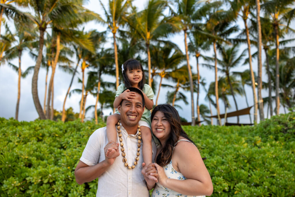 Maui family photography – smiling parents with daughter on dad’s shoulders, palm grove backdrop and soft sunset light near the beach in Maui.