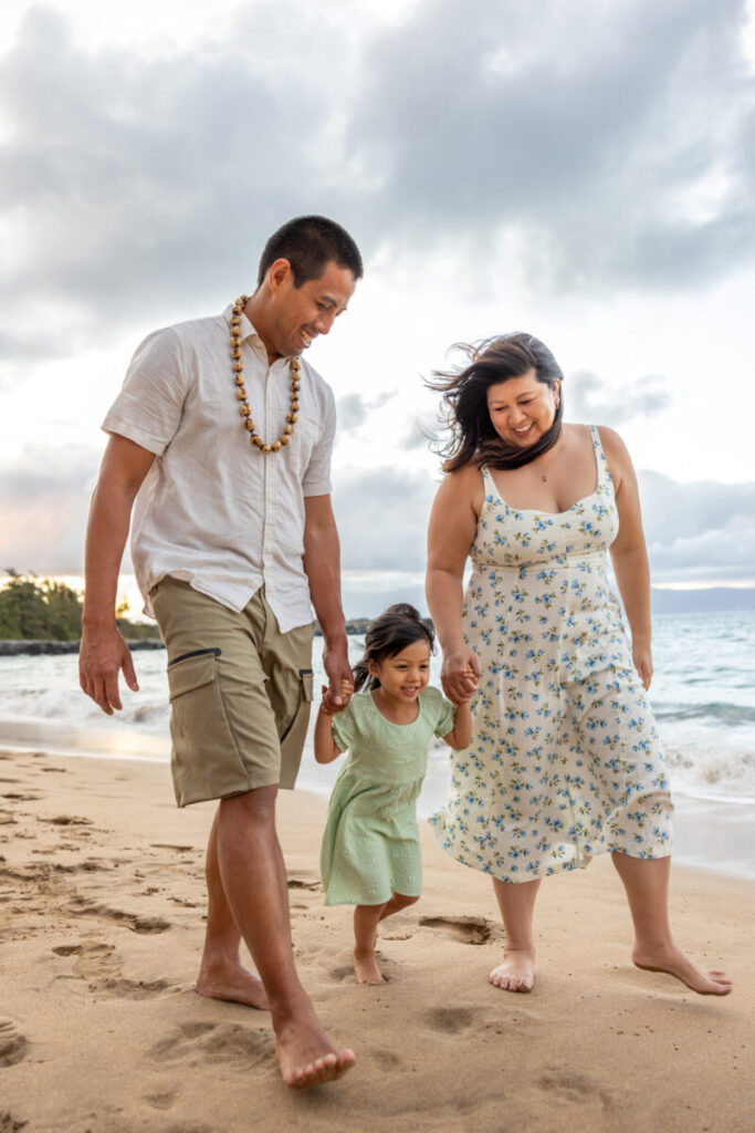 Young girl swinging between her parents’ hands while walking on the sand during sunset Maui family photography at DT Fleming Beach.
