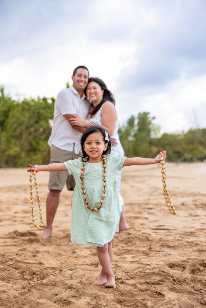Little girl running on the beach holding kukui nut lei with parents in the background during Maui family photography.