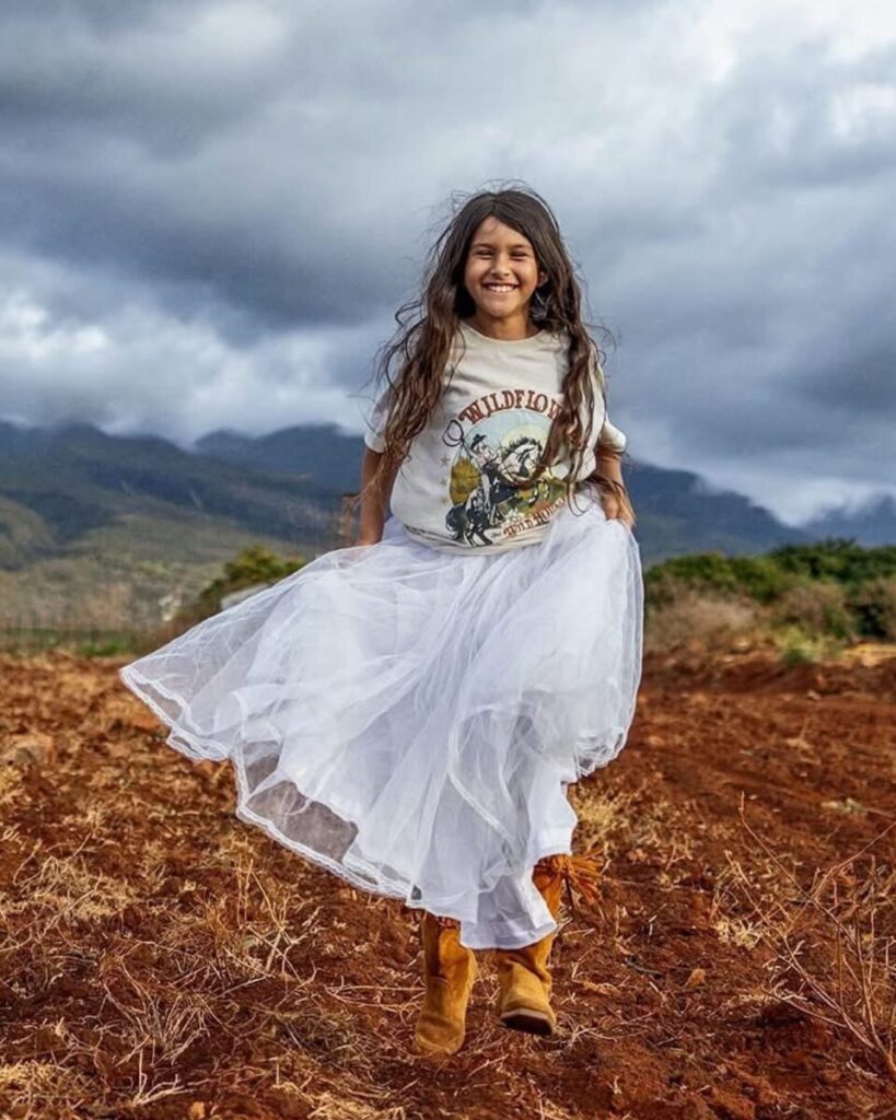 Smiling girl in a white skirt standing in a rustic Maui field with mountains behind her, showcasing one of the Top Maui Photoshoot Locations for natural, earthy portraits.