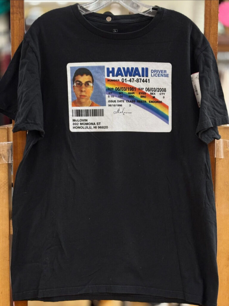 Black T-shirt printed with a vintage Hawaii driver’s license design hanging on a wooden rack at Rainbow Attic