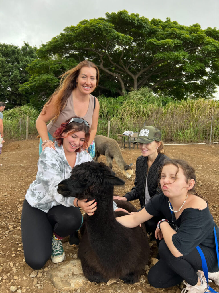 Maui photographers and friends smiling and petting a black alpaca during a farm visit surrounded by green trees at Maui Alpaca