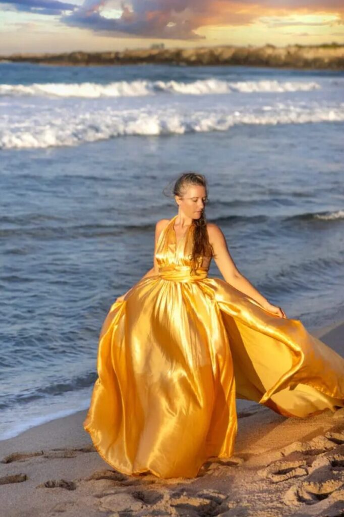 A model wearing a golden Hi Flying Dress walking along a Maui beach, the dress flowing gracefully in the soft shoreline breeze.