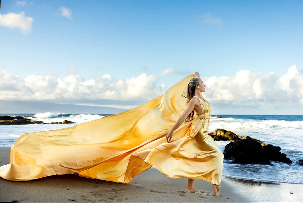 A glamorous Hi Flying Dress photoshoot on Maui featuring a woman in a yellow gown standing on the beach with the sky glowing above.