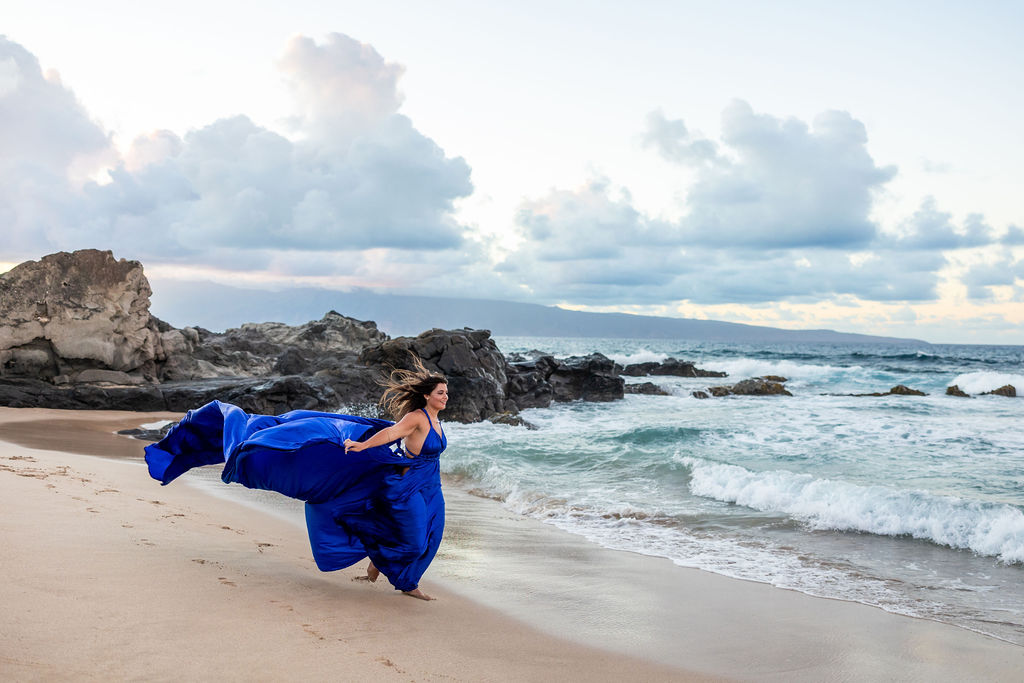 A dramatic Hi Flying Dress photoshoot on Maui, with a woman in a blue gown extending the fabric as ocean waves roll in behind her.
