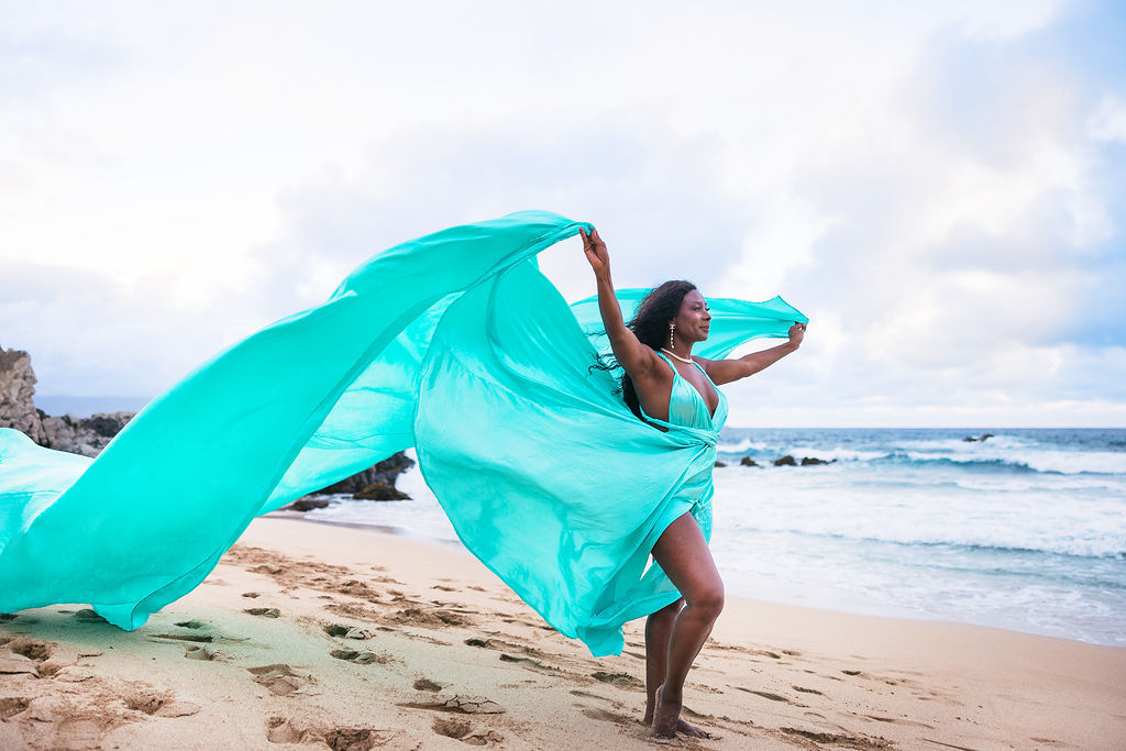 A model in a turquoise Hi Flying Dress standing on golden sand at a Maui beach, with the dress lifting beautifully in the island breeze.