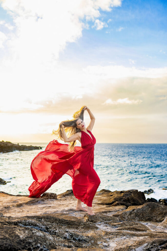 A glowing sunset Hi Flying Dress photoshoot on Maui showing a woman in a red gown with the ocean waves crashing behind her.