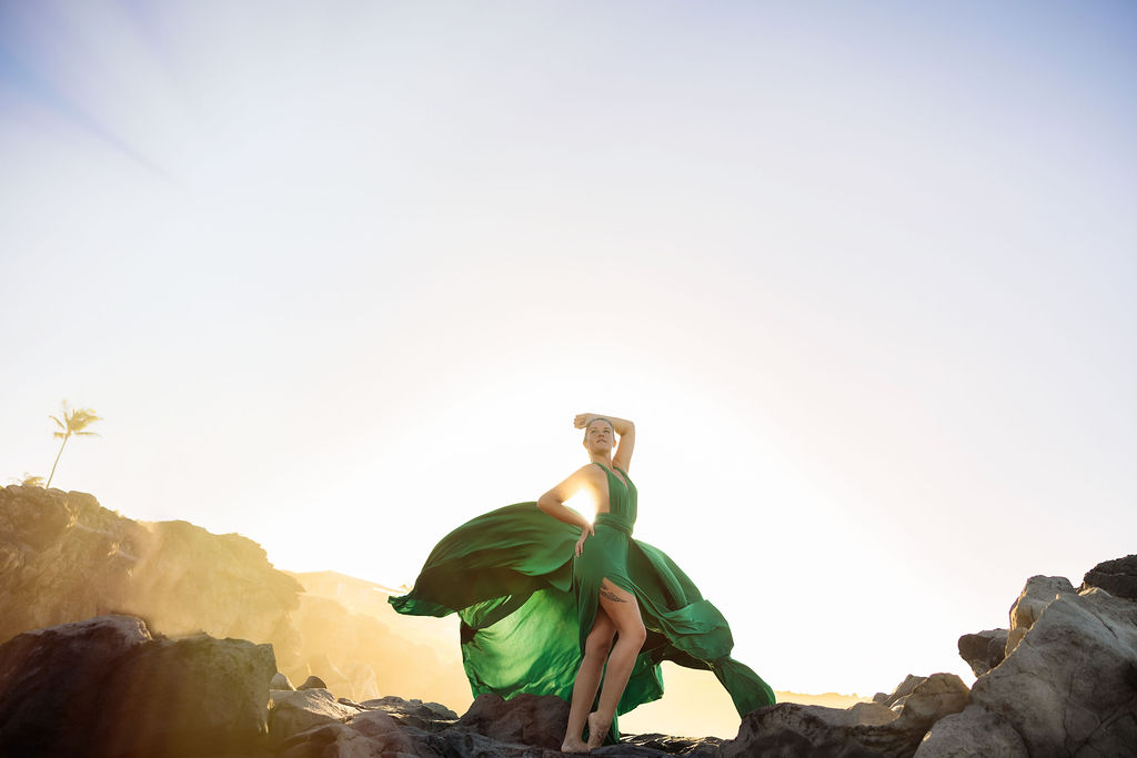 woman in a vibrant green Hi Flying Dress posing on a sunny Maui cliffs, the long fabric floating outward in the wind.