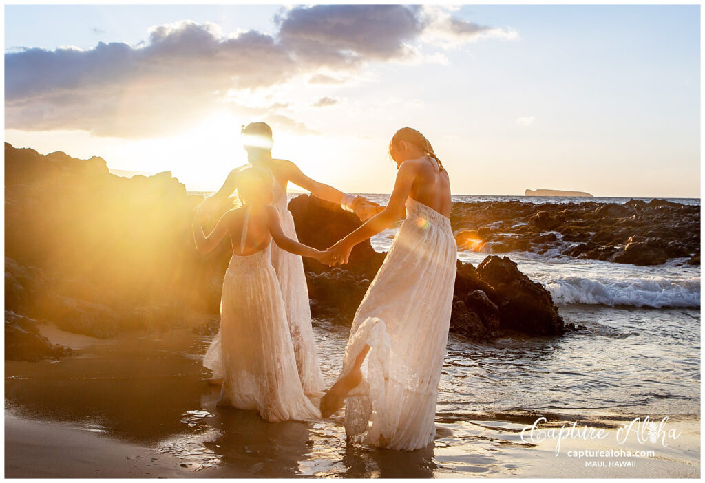 Maui Kids Photography image of three girls holding hands and playing in the shallow ocean at sunset, surrounded by glowing light and rocky Maui coastline.