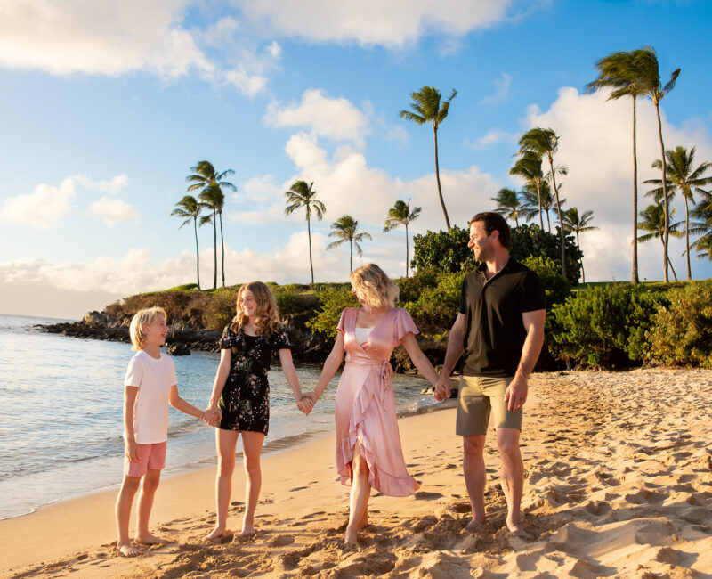 Family of four holding hands, walking on a tropical beach at sunset. Palm trees sway against a blue sky with scattered clouds, creating a joyful atmosphere.