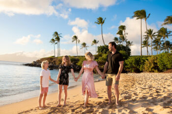 Family of four holding hands, walking on a tropical beach at sunset. Palm trees sway against a blue sky with scattered clouds, creating a joyful atmosphere.