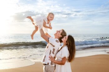A joyful family at the beach, with a man lifting a laughing baby in the air. A woman stands beside them, smiling. The ocean and a blue sky create a serene backdrop.