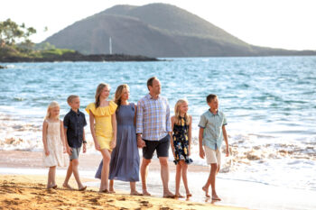 Smiling family of seven walking barefoot along a sandy beach in South Maui during golden hour, with ocean waves and a lush green hill in the background.