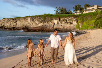 Family of four holding hands walks barefoot on a sandy beach. The two girls wear matching dresses; the parents are in white and beige attire. Sunlit cliffs and ocean waves set a joyful scene.