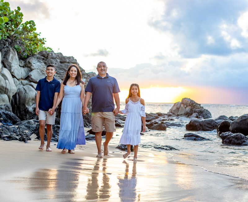 Family of four walks hand in hand on a beach at sunset, surrounded by rocks and greenery, conveying warmth and togetherness.