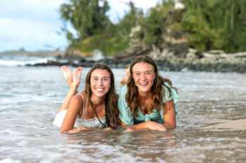 Two young women lie on the wet sand at a beach, smiling at the camera. The ocean waves are gentle, and lush greenery is in the background. The mood is joyful and carefree.