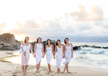 Group of women in matching white dresses walking hand in hand along a rocky beach in South Maui at sunset, with one holding a baby and the ocean waves in the background.
