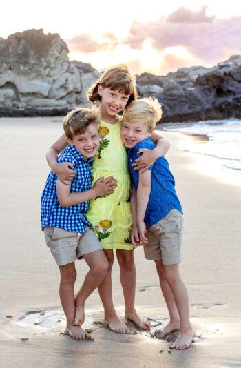 Three smiling children standing barefoot on a sandy beach at sunset, with rocky cliffs in the background. They are hugging and dressed in summer clothes.