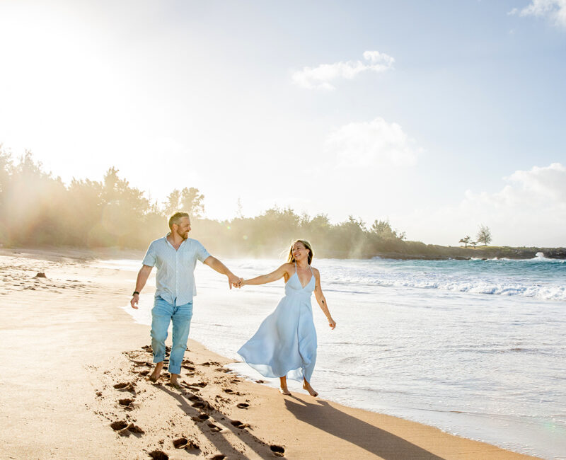 Maui engagement photographer – couple holding hands and walking barefoot along sandy beach with ocean waves.