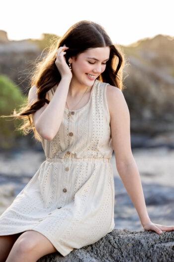 A woman in a light, sleeveless dress sits on a rock, smiling and touching her hair. The background is a soft-focus landscape with rocks and greenery.