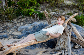 Teen lounges on driftwood by rocky cove, relaxed senior photo, Maui.
