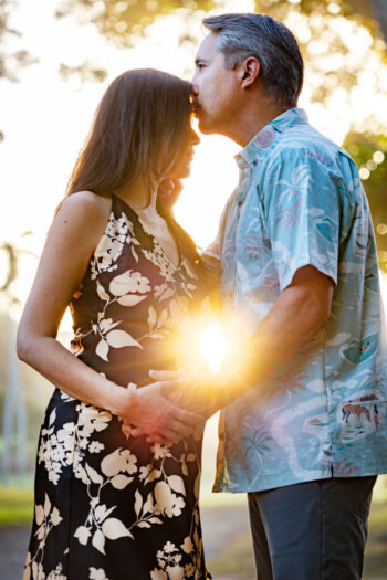 Expecting parents hold belly with warm sun flare between them, Maui.