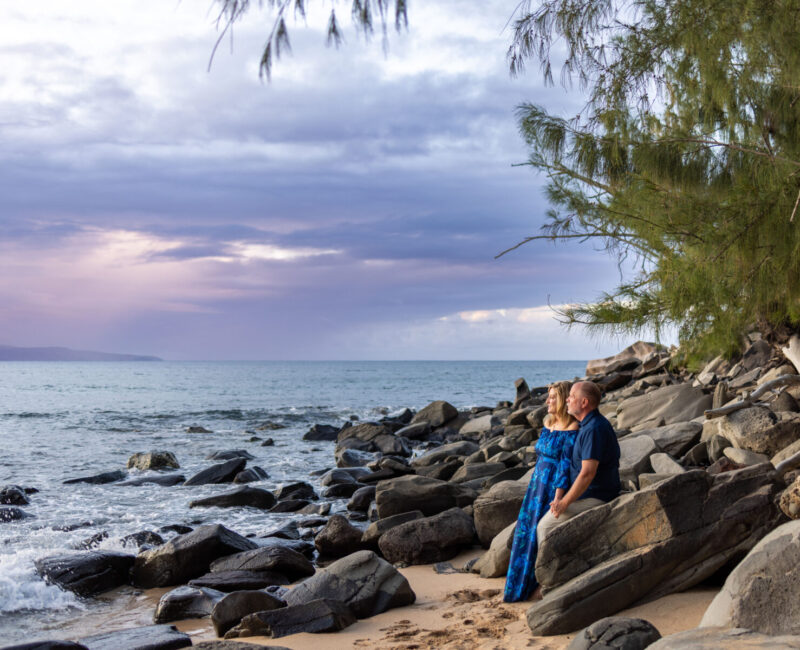 Couple sit side-by-side on rocky shore with warm dusk light, Maui.