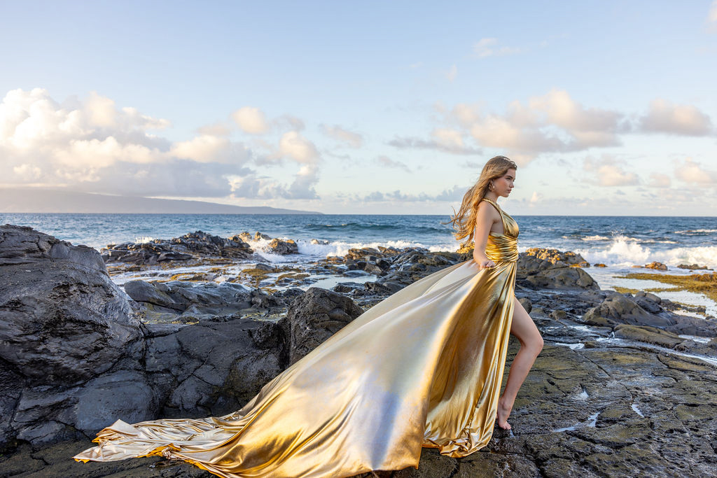 A woman in a flowing, gold flying dress twirling on a Maui beach, with the wind lifting the fabric against a backdrop of turquoise ocean and cliffs