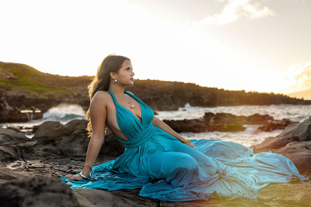 woman sitting on a cliff in a flowing, colorful flying dress twirling on a Maui beach, with the wind lifting the fabric against a backdrop of turquoise ocean 