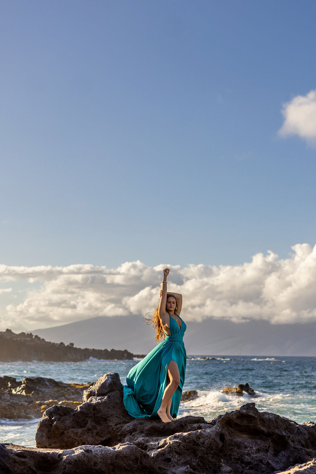 A woman in a flowing, colorful flying dress twirling on a Maui beach, with the wind lifting the fabric against a backdrop of turquoise ocean and cliffs