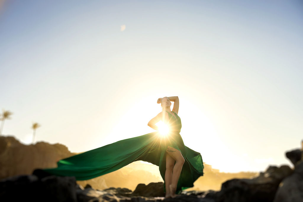 A woman in a flowing, colorful flying dress twirling on a Maui beach, with the wind lifting the fabric against a backdrop of turquoise ocean and cliffs