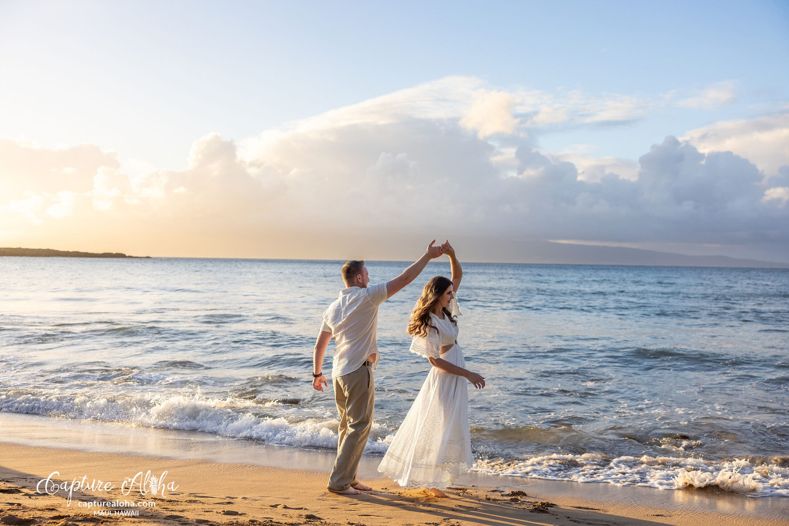 A couple dances on a sandy beach in Maui, with the golden sunset casting a warm glow over the ocean in the background. The woman wears a flowing white dress, and the man is dressed in a light shirt and pants. They smile at each other, holding hands, as gentle waves lap at the shore. The sky is a mix of soft pinks, oranges, and purples.