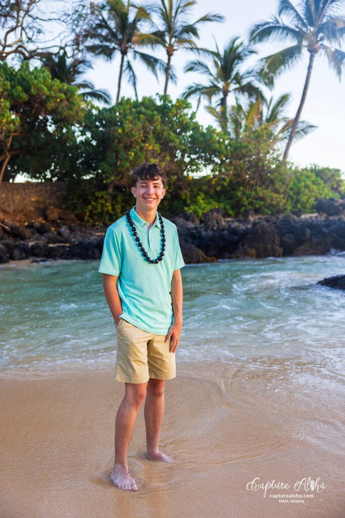 Senior portrait of a student in Maui, standing on a scenic beach with lush greenery and the ocean in the background. The student is dressed in a casual yet stylish outfit, smiling confidently while the warm golden light of sunset creates a soft glow. The vibrant blue sky and gentle waves enhance the tropical atmosphere, showcasing the beauty of the Hawaiian landscape