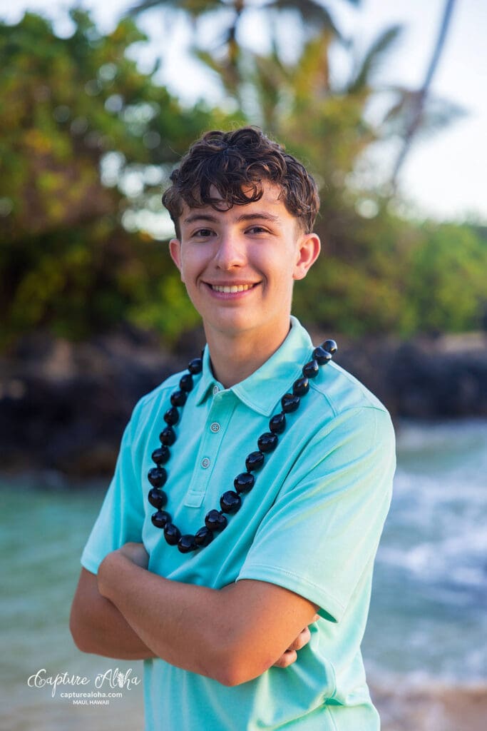 Senior portrait of a student in Maui, standing on a scenic beach with the turquoise ocean stretching into the distance. The student, wearing a light, flowing outfit, smiles confidently while the golden hour light bathes the scene in warmth. The soft breeze tousles their hair, and the sky above is a perfect blend of pink, orange, and blue hues as the sun sets behind the distant mountains. Lush tropical greenery borders the beach, and the gentle waves lap at the shore, capturing the serene beauty and vibrant atmosphere of Maui