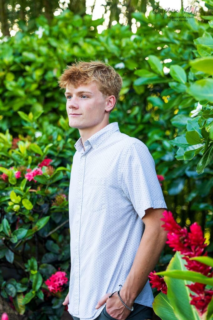 Senior portrait of a student standing amidst lush tropical greenery on a Maui hillside, with vibrant foliage framing the scene. The student, dressed in a casual yet stylish outfit, smiles softly while the sunlight filters through the leaves, casting a warm glow on their face. The rich green plants and trees create a natural, serene backdrop, with hints of colorful flowers adding pops of color. The peaceful atmosphere of the Hawaiian landscape is enhanced by the soft light and the distant hum of nature, capturing the essence of Maui's beauty and tranquility