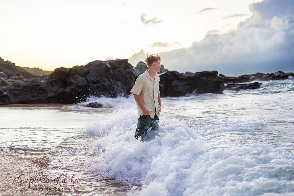 Senior portrait of a student standing in the shallow waves of a Maui beach, with the ocean gently lapping at their feet. The student, dressed in a light, flowing outfit, smiles with a sense of joy and confidence as the soft waves roll around them. The golden glow of the setting sun casts a warm light across the scene, reflecting off the water and creating a dreamy, ethereal atmosphere. In the background, the horizon stretches into a soft gradient of pinks and oranges, while the lush green coastline and distant mountains complete the serene, tropical backdrop