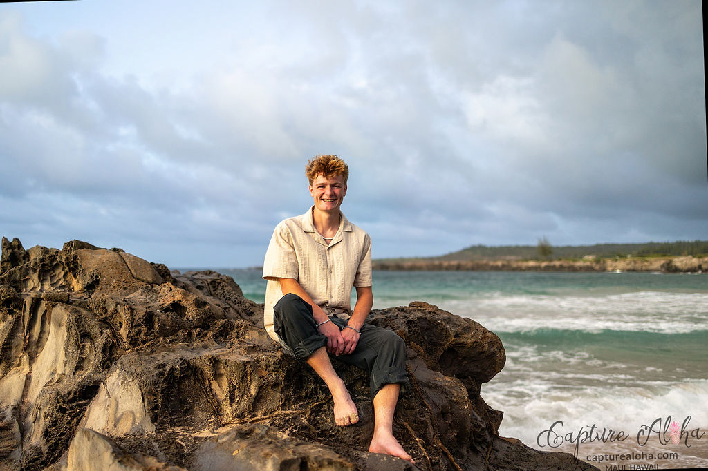 Senior portrait of a student standing on rugged rocks along a Maui coastline, with the ocean crashing gently below. The student, dressed in a casual yet chic outfit, gazes confidently into the distance, bathed in the warm, golden light of the setting sun. The dramatic landscape of black volcanic rocks contrasts with the vibrant blues of the ocean and the soft pastels of the sunset sky. The student’s relaxed pose and the serene backdrop of swaying palm trees and distant mountains highlight the natural beauty and peaceful energy of Maui