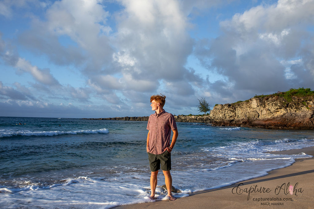 Senior portrait of a student standing on a Maui beach at sunset, with the golden light casting a warm glow over the scene. The student smiles softly, dressed in a flowing, light-colored outfit that catches the breeze. The ocean stretches out behind them, its turquoise waters blending into the horizon where the sky is painted in hues of pink, orange, and purple. Tall palm trees gently sway, and the soft waves create a calming rhythm, highlighting the natural beauty and serene atmosphere of the Hawaiian landscape