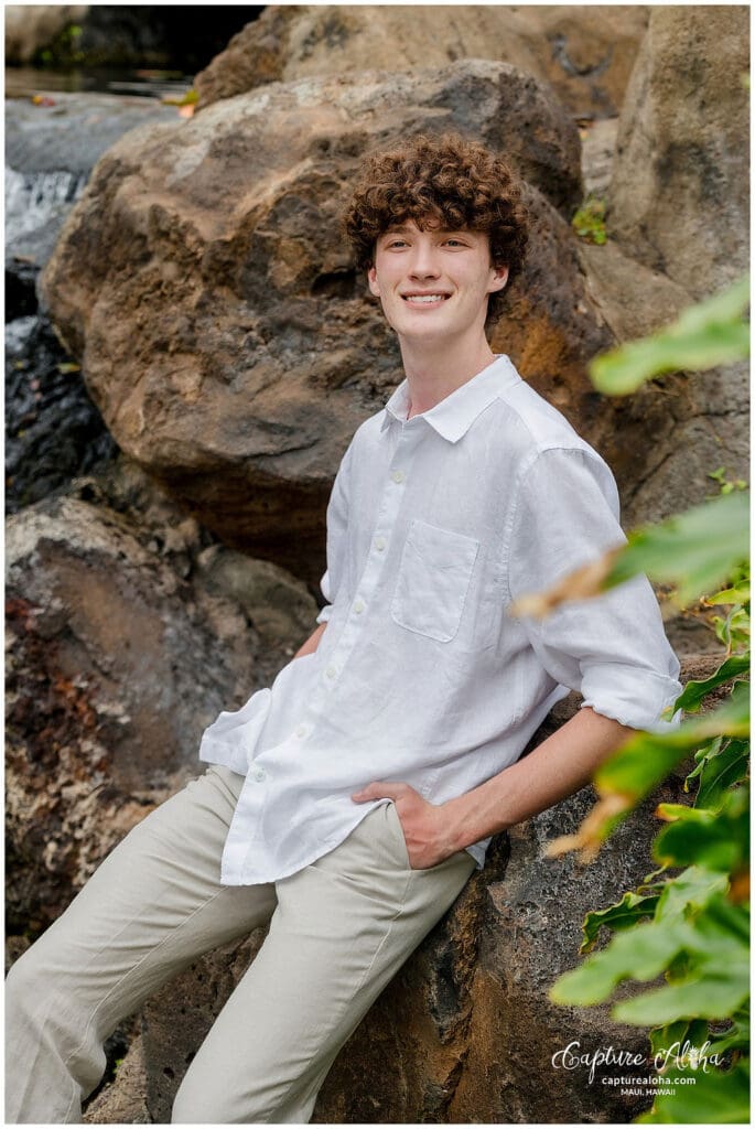 Senior portrait of a Maui student, standing confidently on a rocky beach with the ocean and mountains in the background. The warm, golden sunset light casts a soft glow, highlighting the student’s smile and vibrant attire. The scene captures the natural beauty of Maui and the student’s sense of pride and achievement