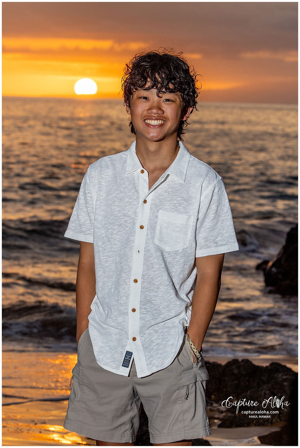 Senior portrait of a student on a Maui beach, with a breathtaking backdrop of the Pacific Ocean and towering mountains. The student, dressed in a casual yet elegant outfit, stands with a relaxed pose, a warm smile on their face as the soft glow of the setting sun illuminates them. The vibrant colors of the sky — shades of pink, purple, and orange — create a stunning contrast against the deep blue of the ocean. Lush tropical plants frame the scene, while gentle waves roll onto the shore, embodying the peaceful and idyllic atmosphere of Maui