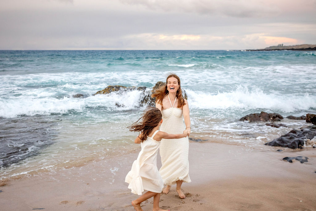 Mother and daughter dancing barefoot on Maui Beach during a family portrait session, smiling and enjoying the sunset.