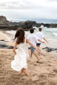 Siblings laughing and playing in the sand while their parents watch with joy during a Maui beach photoshoot.